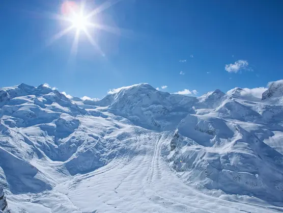 Ein atemberaubender Blick auf schneebedeckte Berge unter strahlend blauem Himmel. Die Sonne scheint hell, beleuchtet die Gipfel und schafft eine ruhige, klare Winterlandschaft. Im Hintergrund schweben flauschige Wolken.