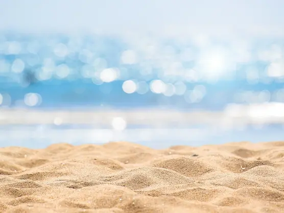 Nahaufnahme eines Sandstrandes mit sonnenbeschienenen Meereswellen im Hintergrund. Das Wasser glitzert im hellen Sonnenlicht und erzeugt einen Bokeh-Effekt. Der Sand im Vordergrund ist glatt und sonnenbeschienen und fängt eine ruhige Küstenszene ein.